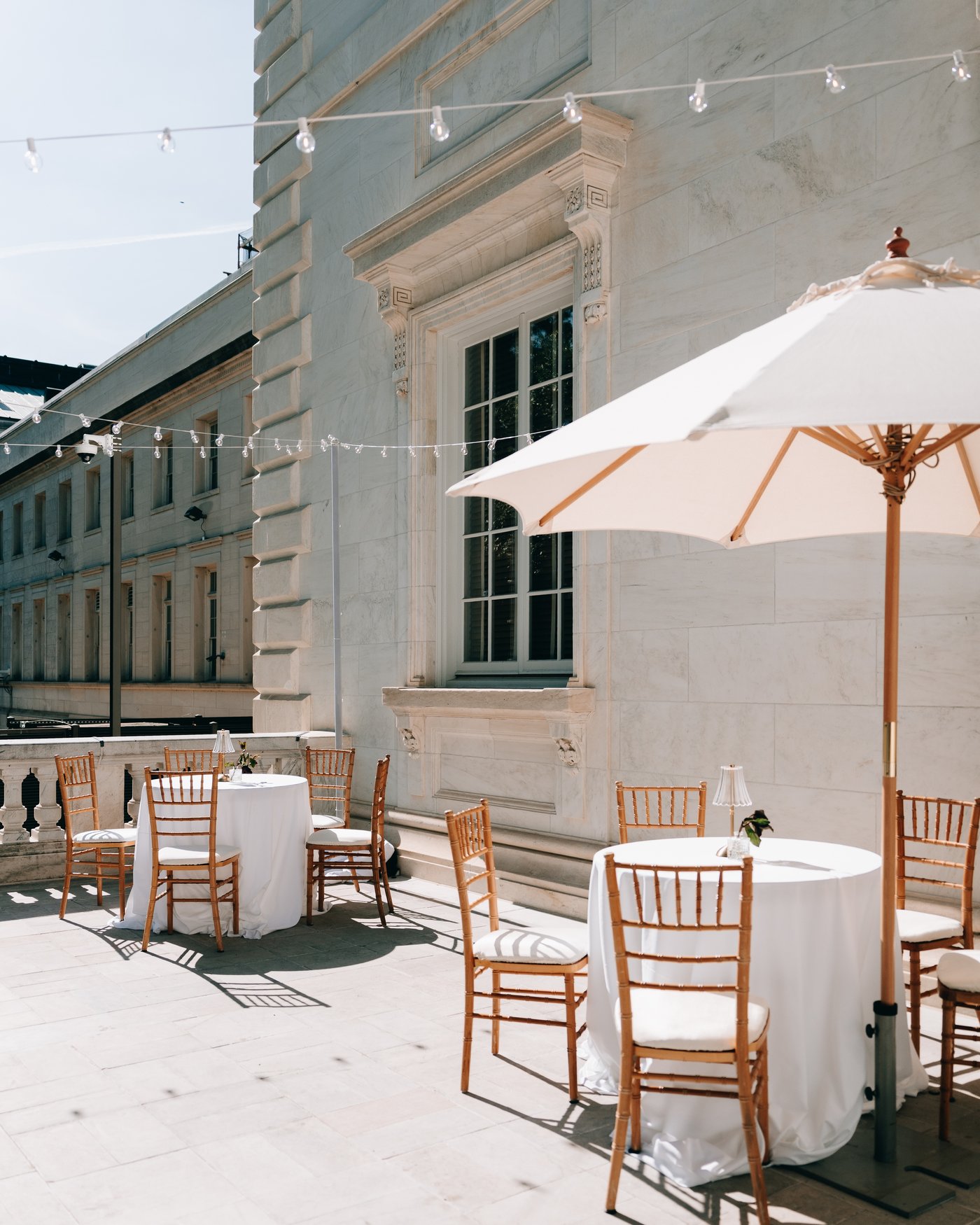 The terrace before anyone arrives: cocktail tables, gilt chairs and a canvas umbrella under strung lights