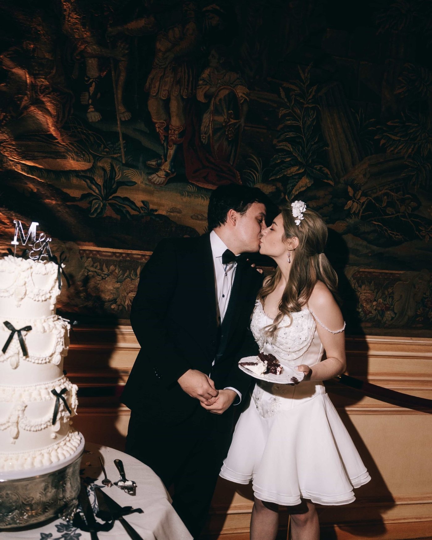 A couple kissing beside a tall tiered cake tied with black bows, a painted mural behind them