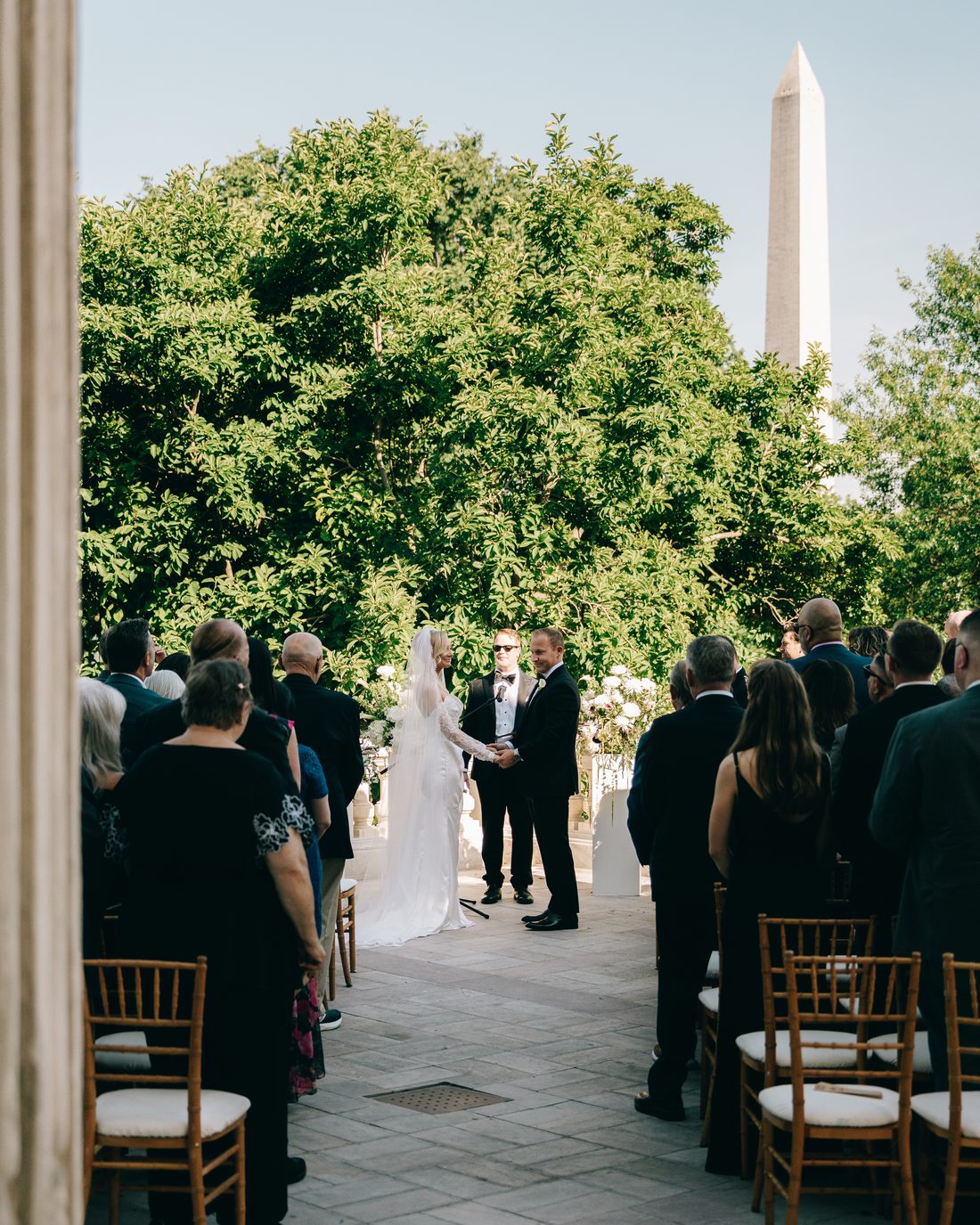 Guests standing for the ceremony, framed by a tall column, the couple small at the front