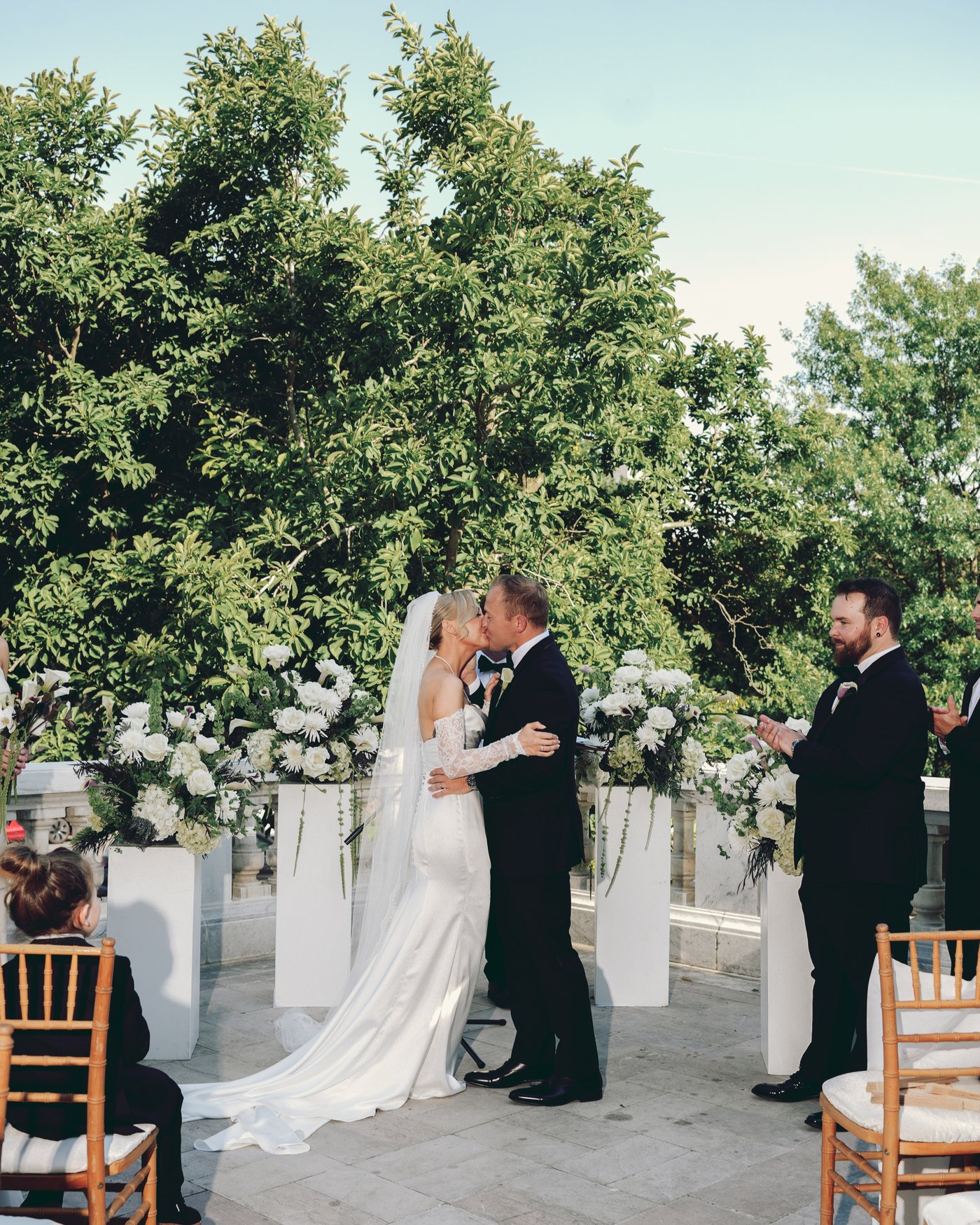 The first kiss under a flowered arch, the officiant beside them and the guests seated on the grass