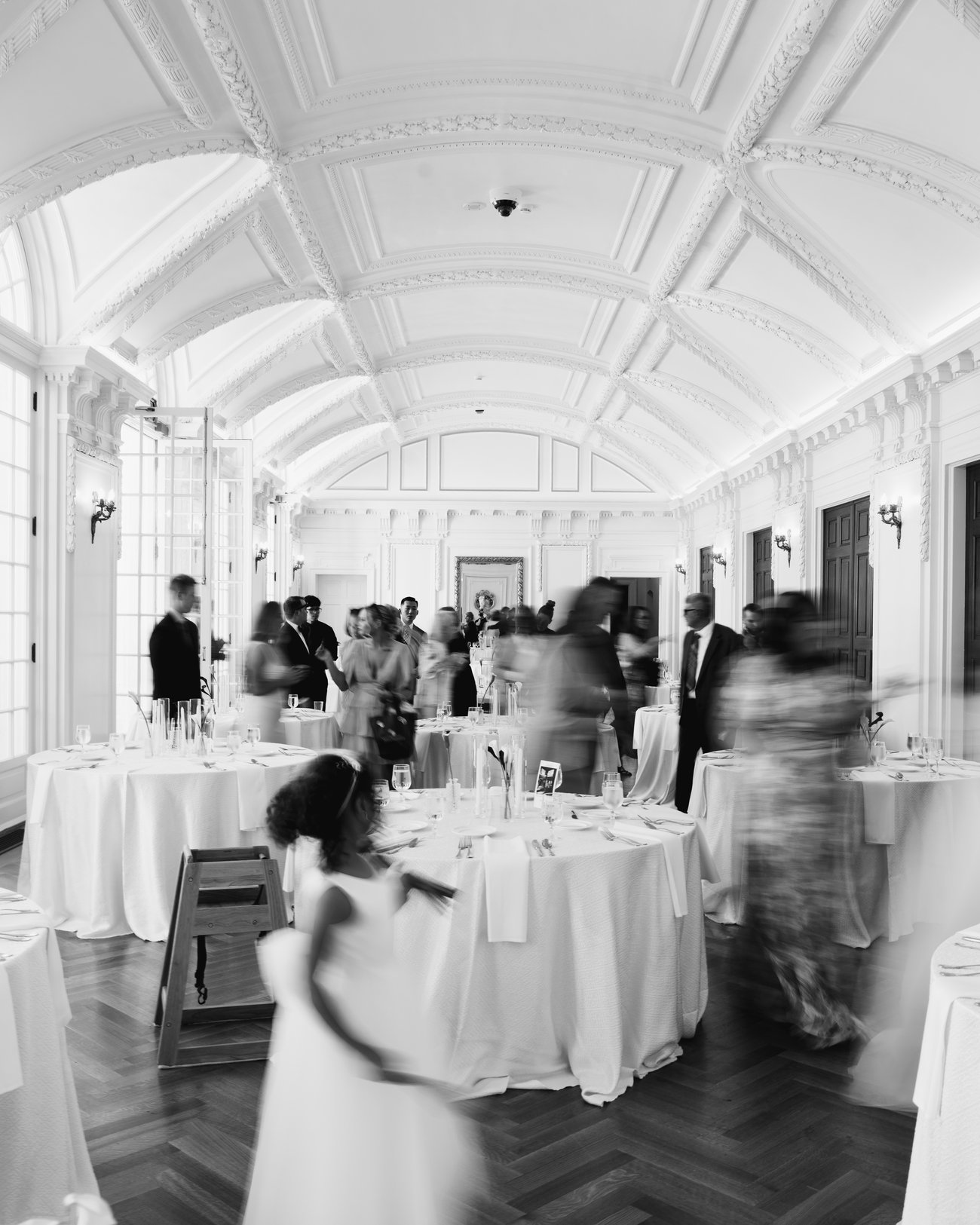 Black and white, a long exposure of the reception, the guests blurred between the tables under a vaulted ceiling