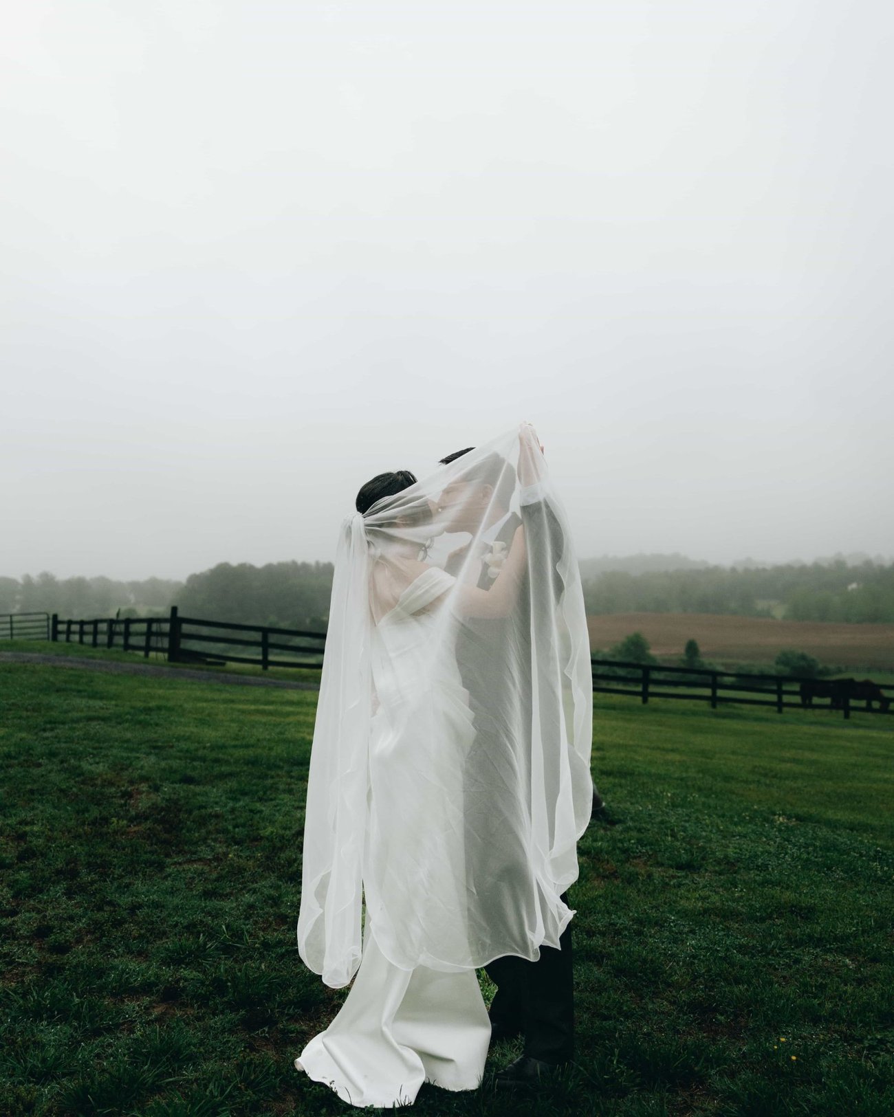 A bride holding her veil up over both of them, kissing the groom underneath it in a misty pasture
