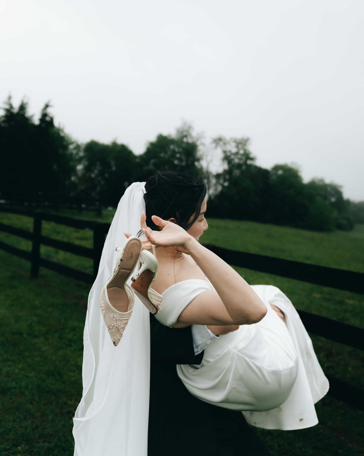 A groom carrying the bride across wet grass, her beaded heels dangling from her hand