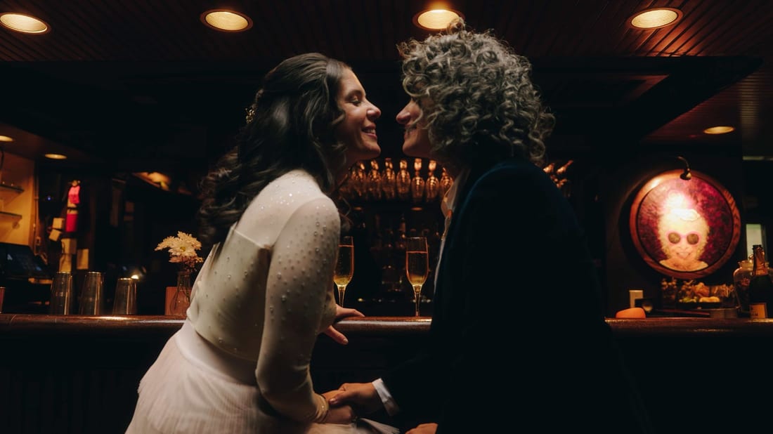 Two brides at a dark bar, faces almost touching, two glasses of champagne waiting behind them