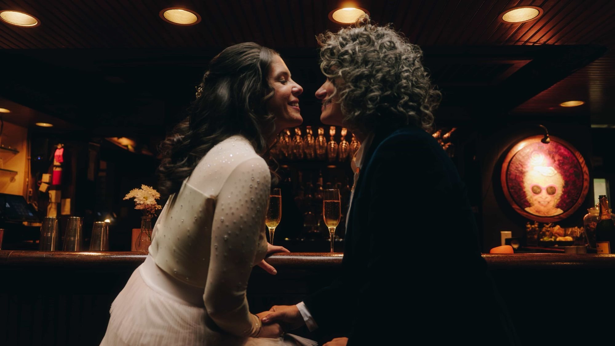 Two brides at a dark bar, faces almost touching, two glasses of champagne waiting behind them