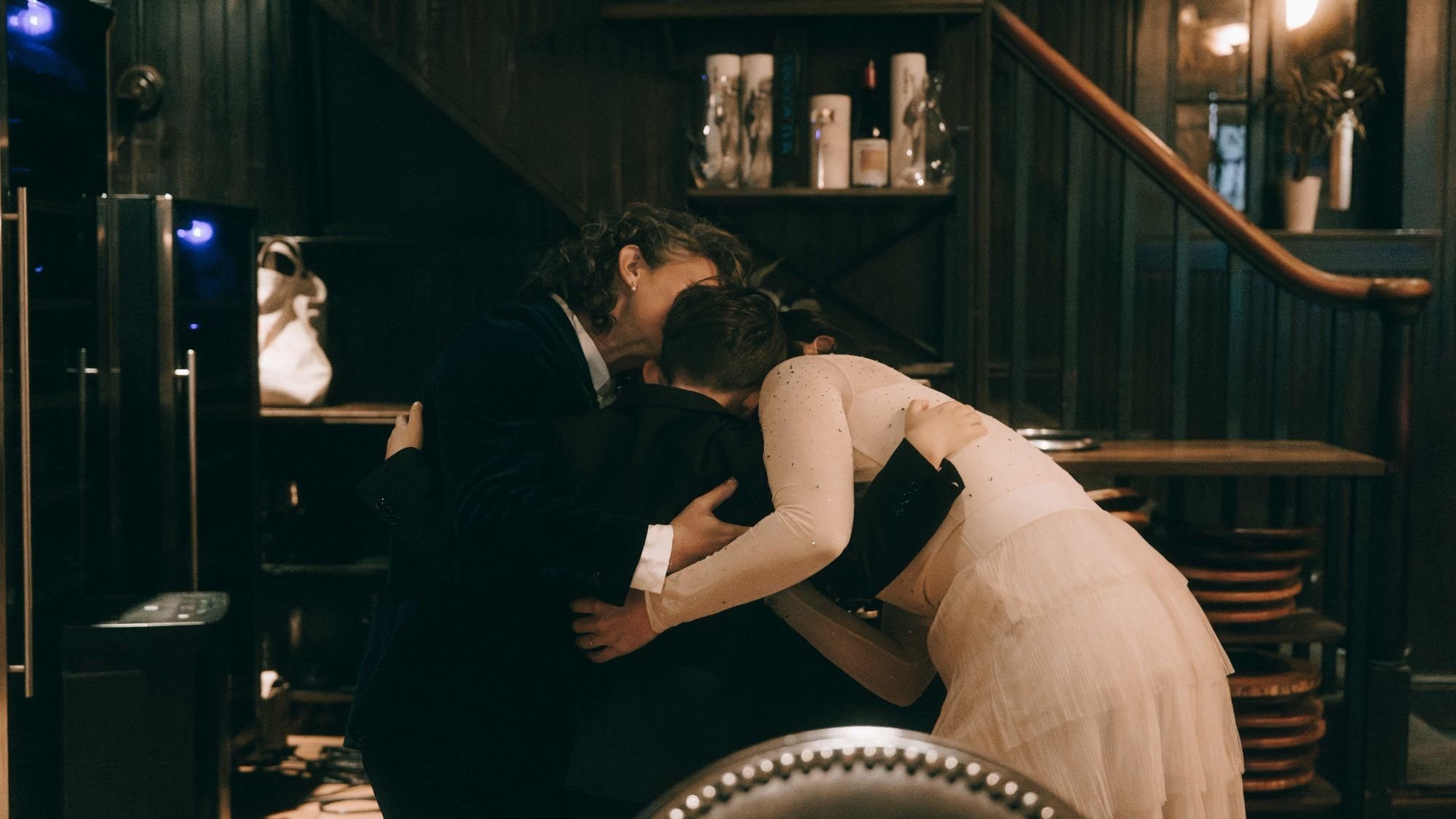 Three people folded into one embrace at the foot of a dark staircase, a pale tulle skirt spilling out of it