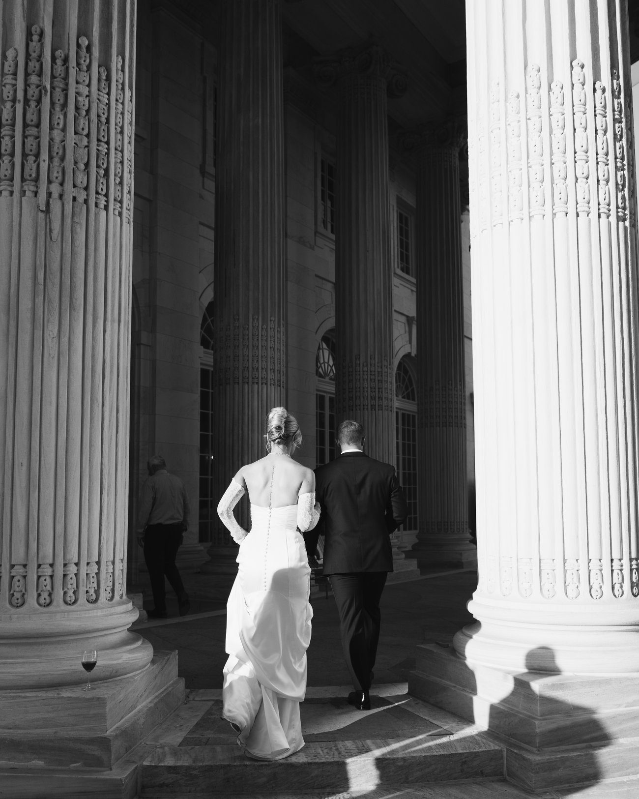 Black and white, a bride gathering her skirt as she walks with the groom between enormous fluted columns