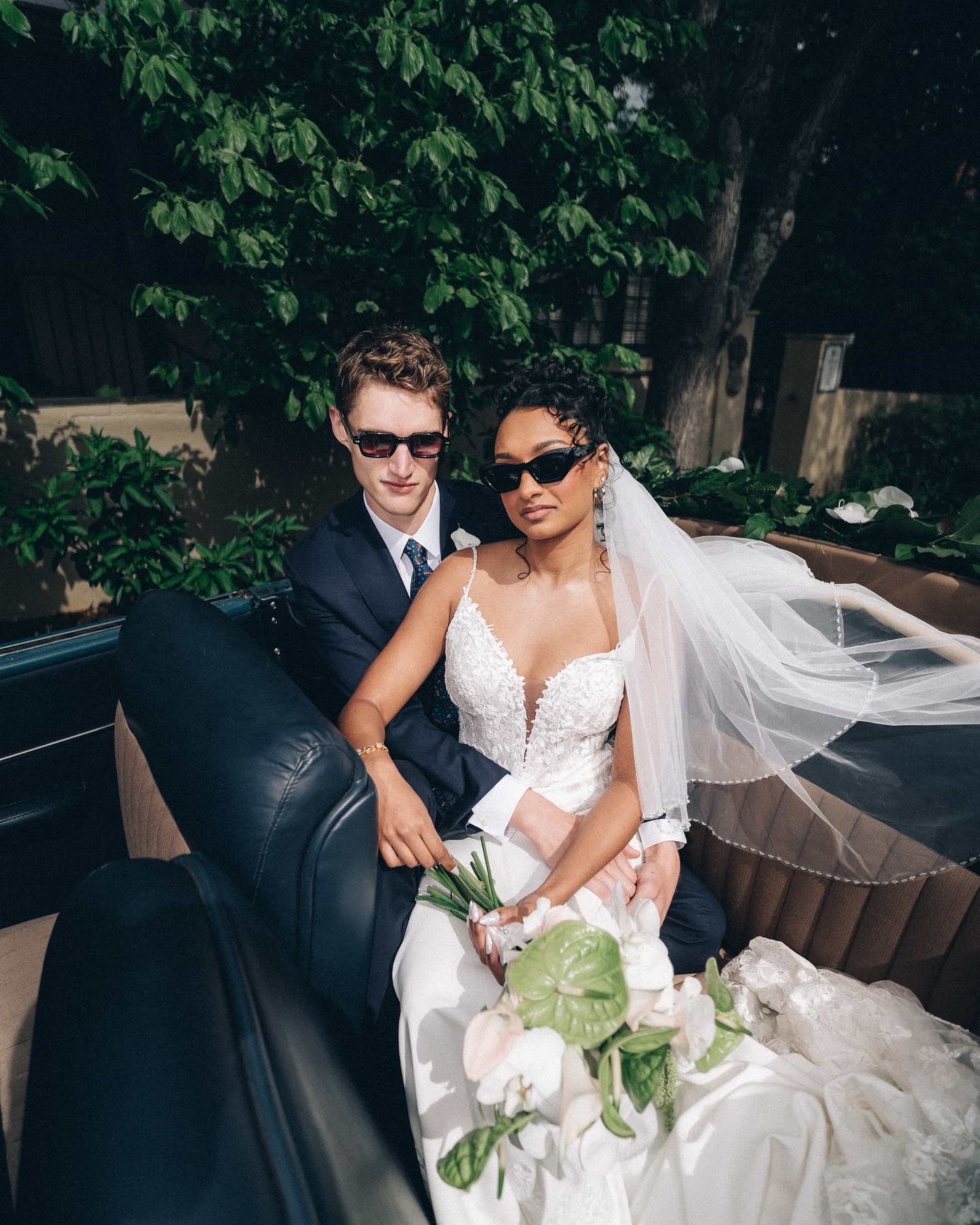 A couple in sunglasses riding in the back of an open car, the veil spilling over the door and lilies in her lap