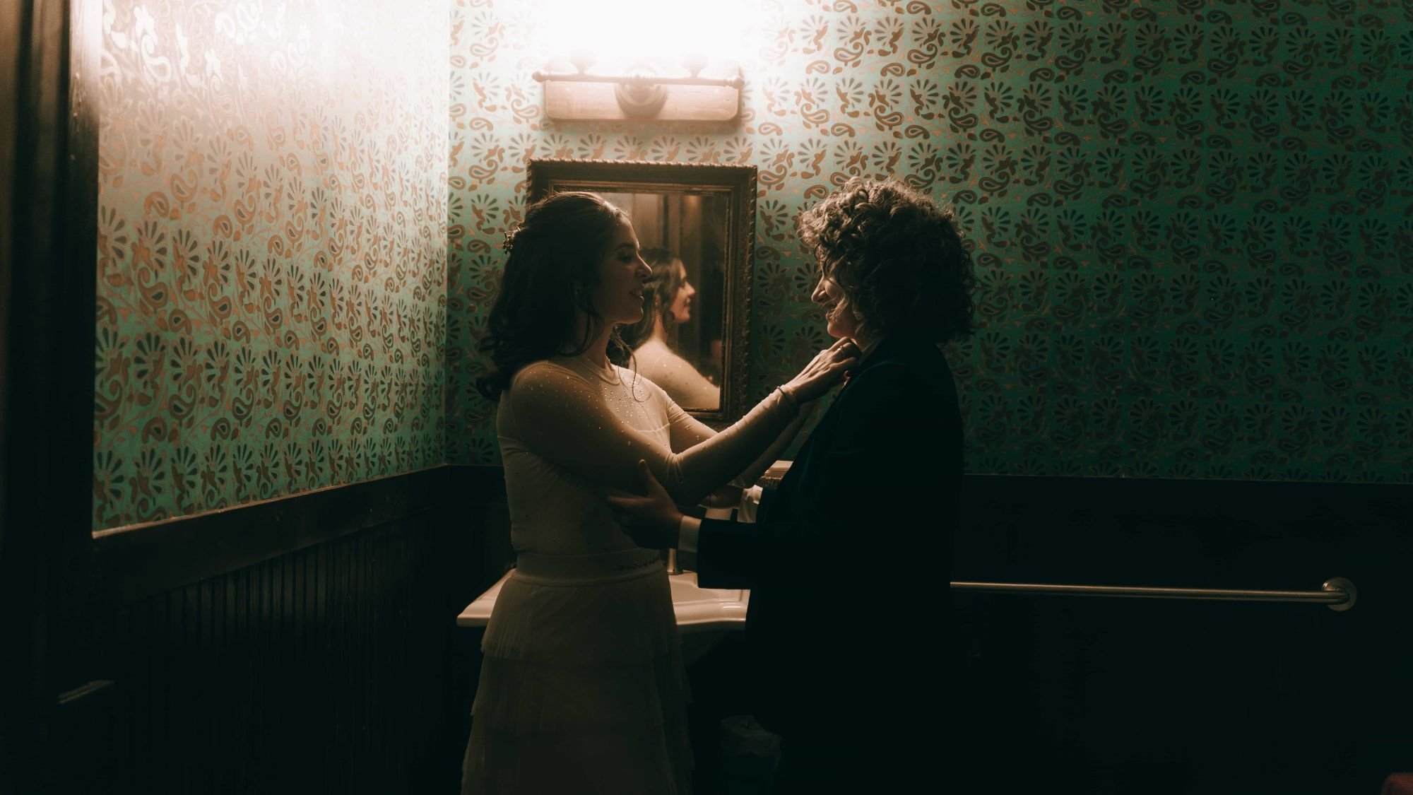 Two brides face to face in a papered washroom, one reaching up to straighten the other's collar