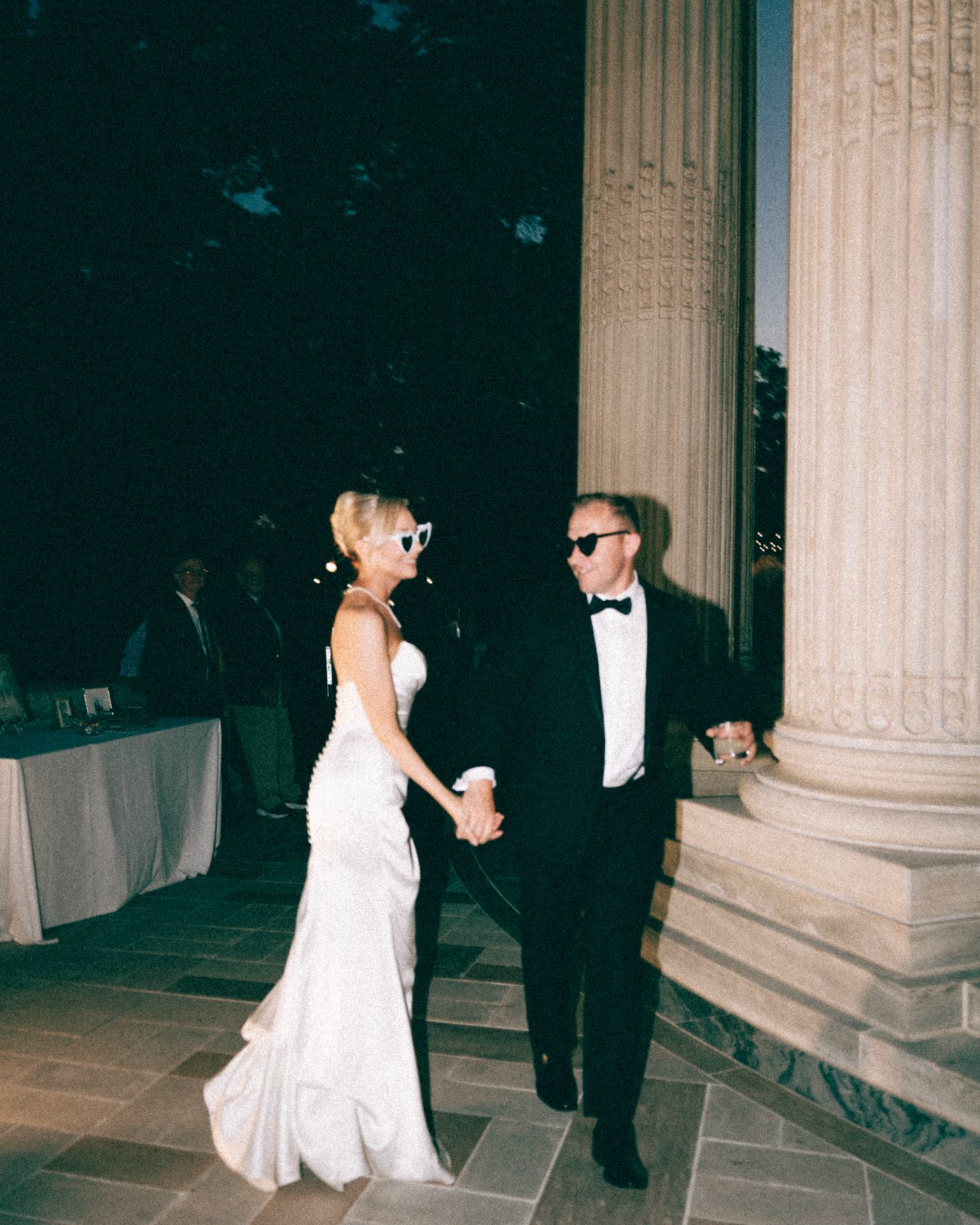 A couple in sunglasses walking a flagstone terrace at dusk, their guests trailing behind them between the columns
