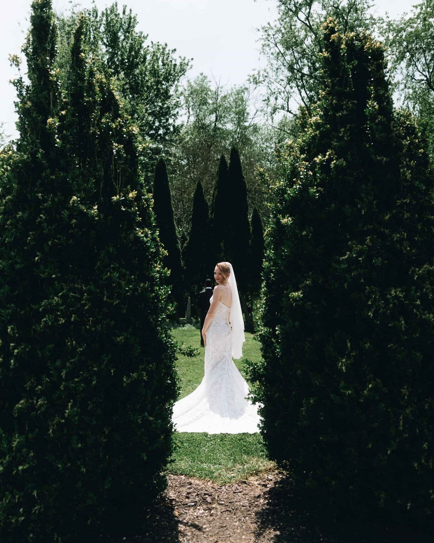 A bride standing alone at the end of a corridor of tall clipped hedges