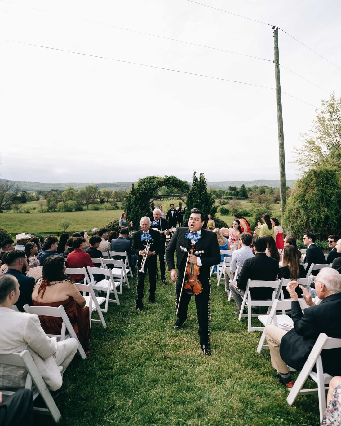 Mariachi musicians singing their way down the grass aisle of a hilltop ceremony, guests turned in their chairs to watch