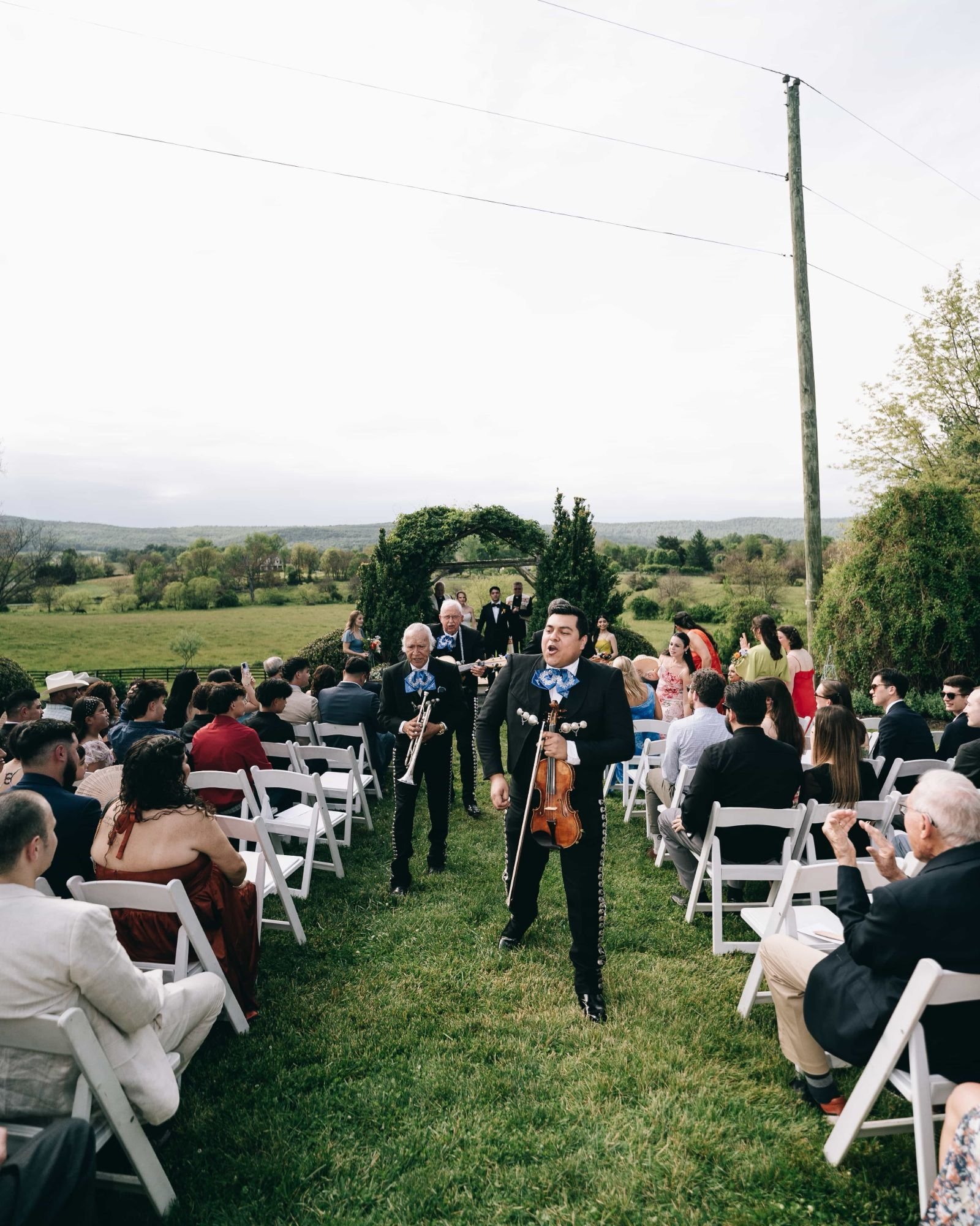 Mariachi musicians singing their way down the grass aisle of a hilltop ceremony, guests turned in their chairs to watch