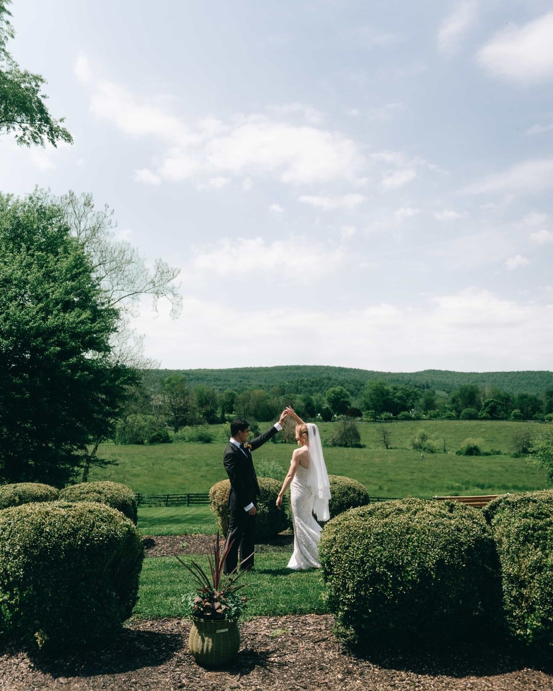 A couple alone on a lawn between clipped boxwoods, a wide green valley and a bigger sky behind them