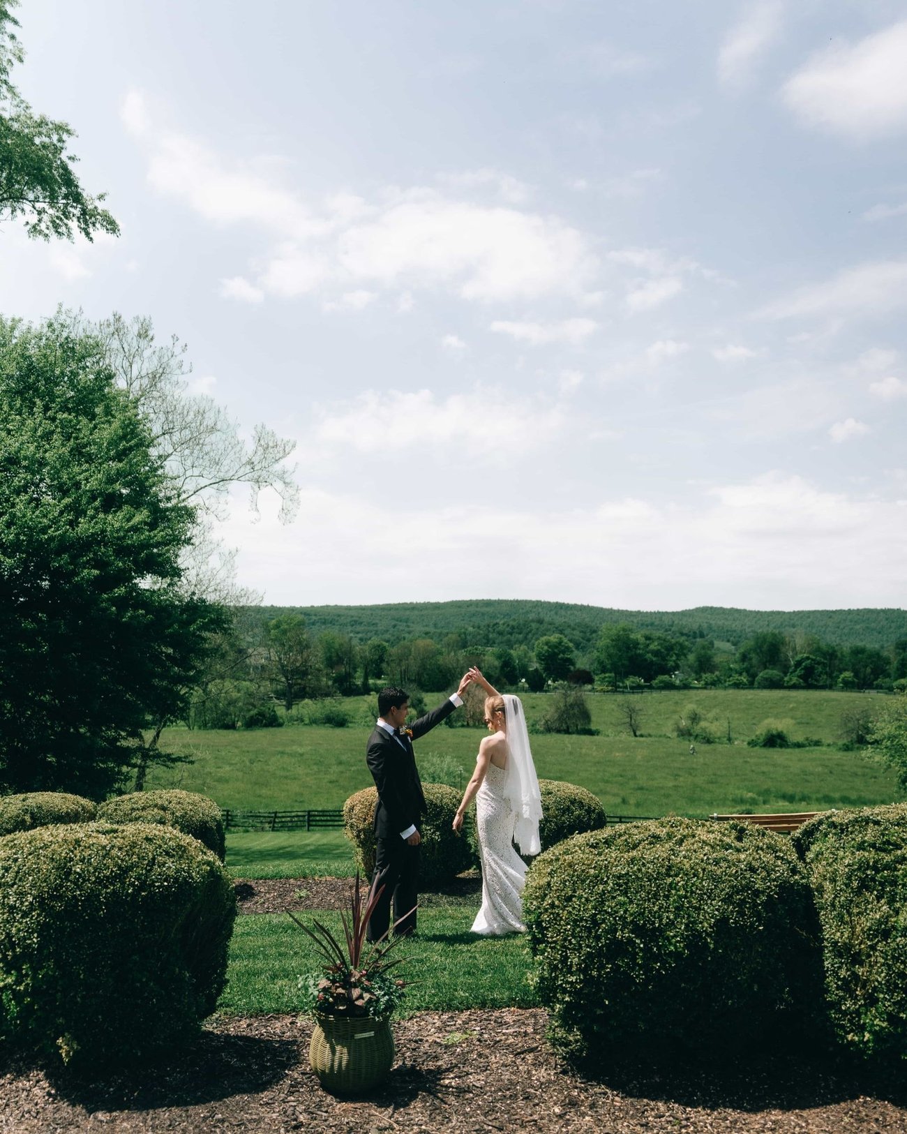 A couple alone on a lawn between clipped boxwoods, a wide green valley and a bigger sky behind them