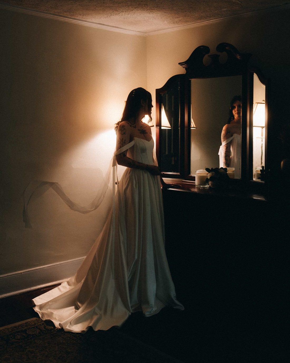 A bride alone at a mirror in lamplight, the train of her gown running out behind her