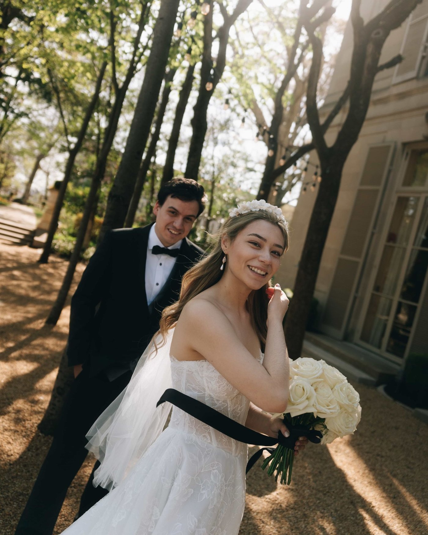 A bride laughing in dappled woodland light, white roses in her gloved hand and the groom close behind her