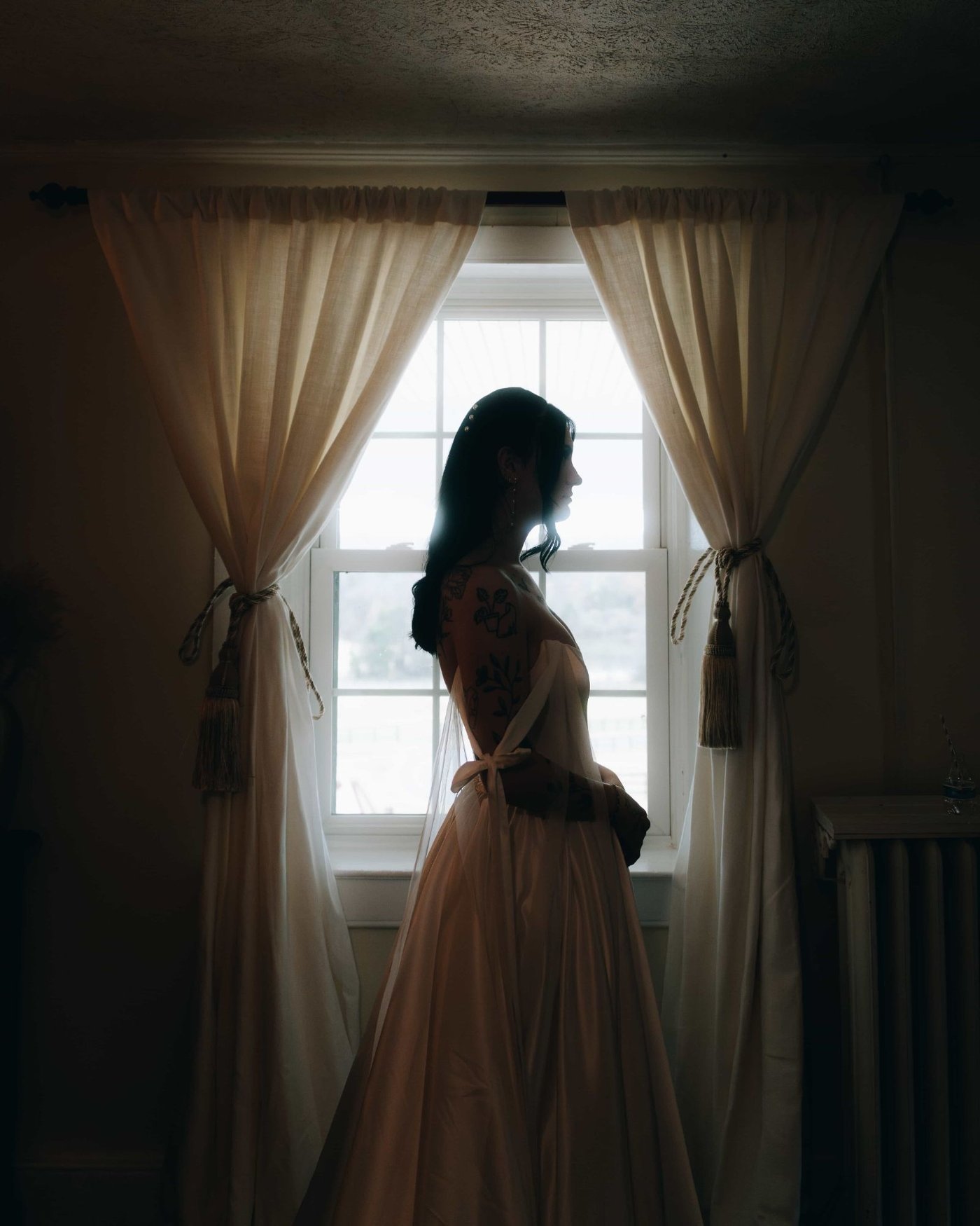 A bride in profile at a curtained window, the light coming through the embroidery down the back of her gown
