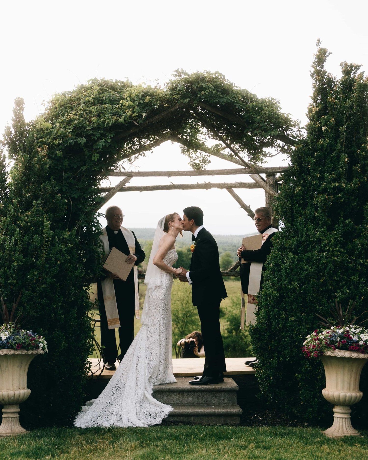 A ceremony under a leafy arbour between hedges, the couple kissing with the officiant beside them
