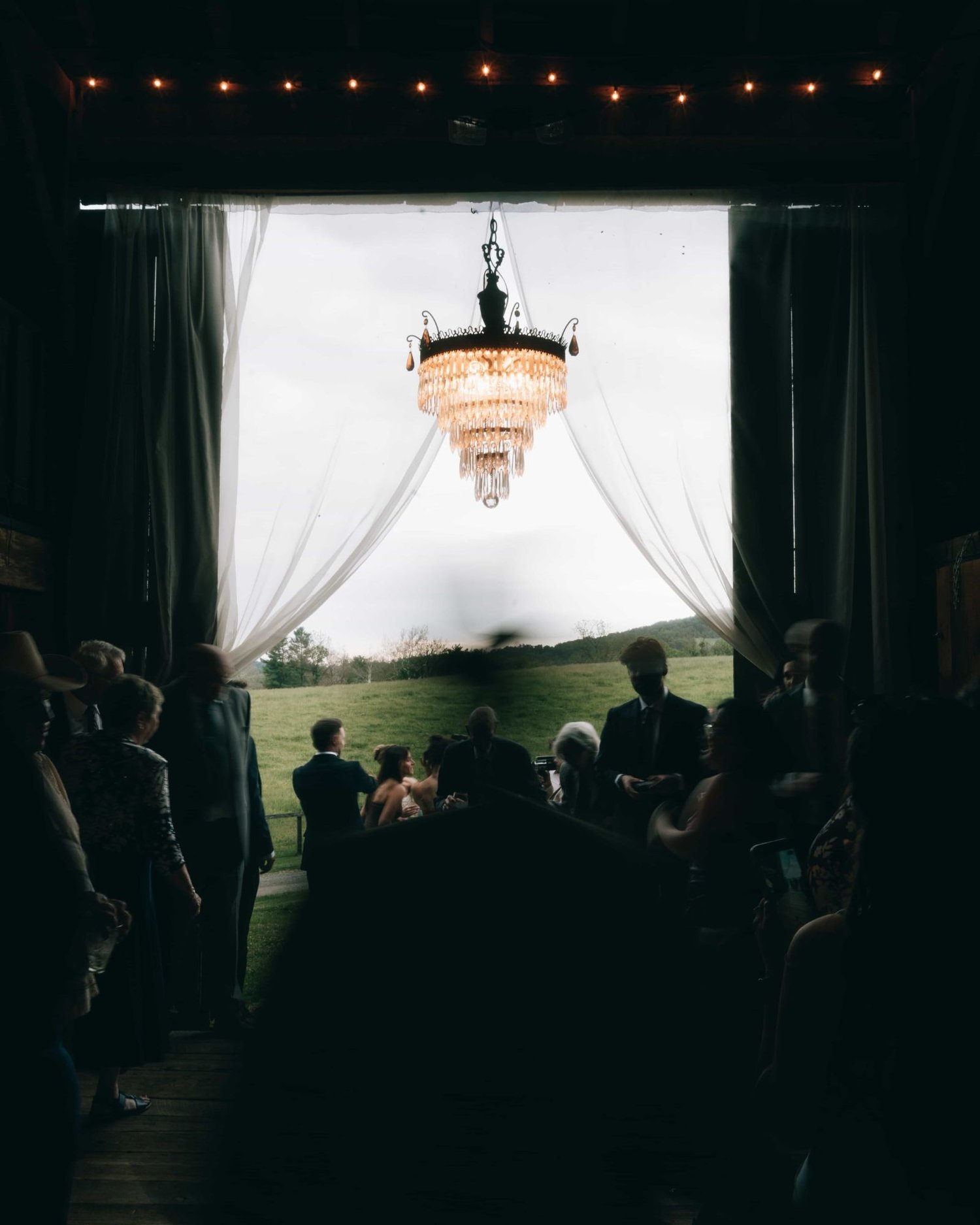 Looking down a dark aisle toward a bright draped opening with a chandelier hanging in it, the guests in silhouette