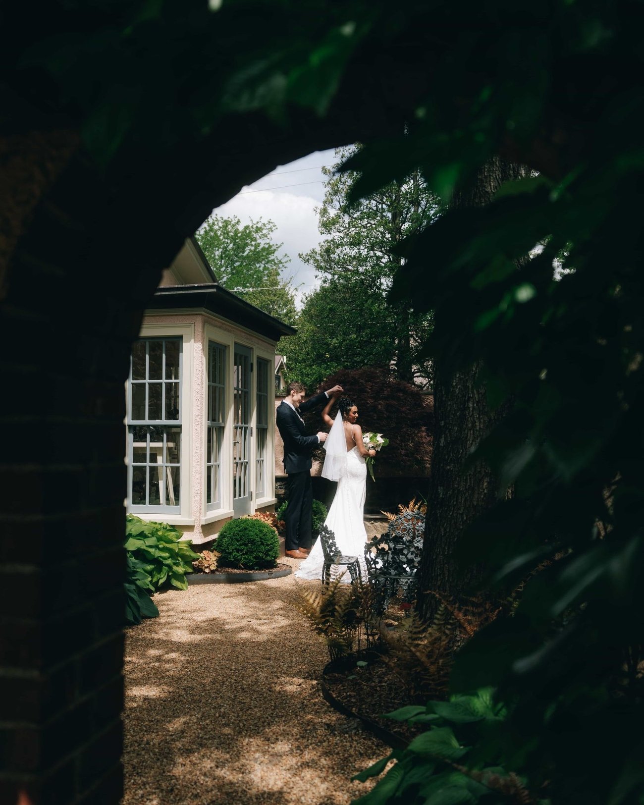 A couple on a garden path beside a white conservatory, seen through an arch of dark leaves