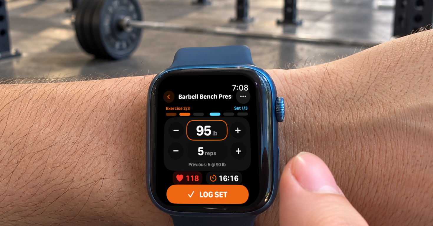 Apple Watch on a lifter's wrist showing Massiv's set screen for a barbell bench press at 95 lb for five reps, with Previous: 5 @ 90 lb and a Log Set button, in front of a loaded barbell
