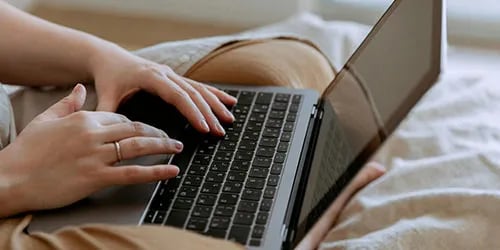 A white woman typing on her laptop in a comfy blanket. 