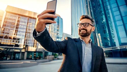 A business person taking a selfie with their smart phone in front of a commercial building.