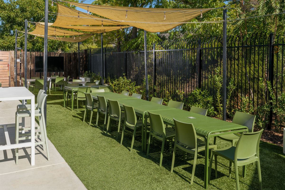 Long tables set for a group under shade sails in the Back Garden, with an outdoor TV on the fence