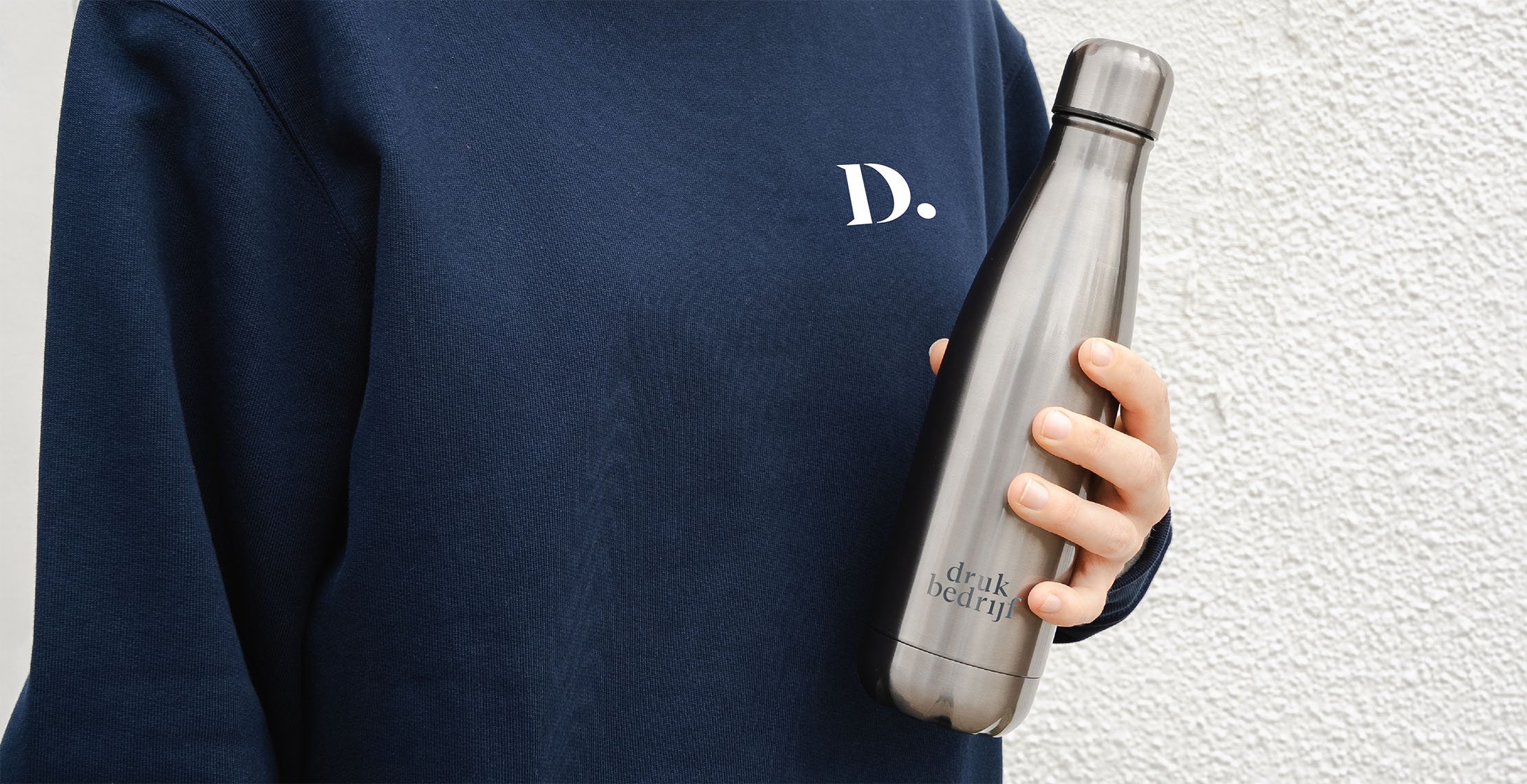 Bedrukte relatiegeschenken voor op werk met een bedrukte trui en een gegrafeerde drinkfles in de hand voor witte muur.