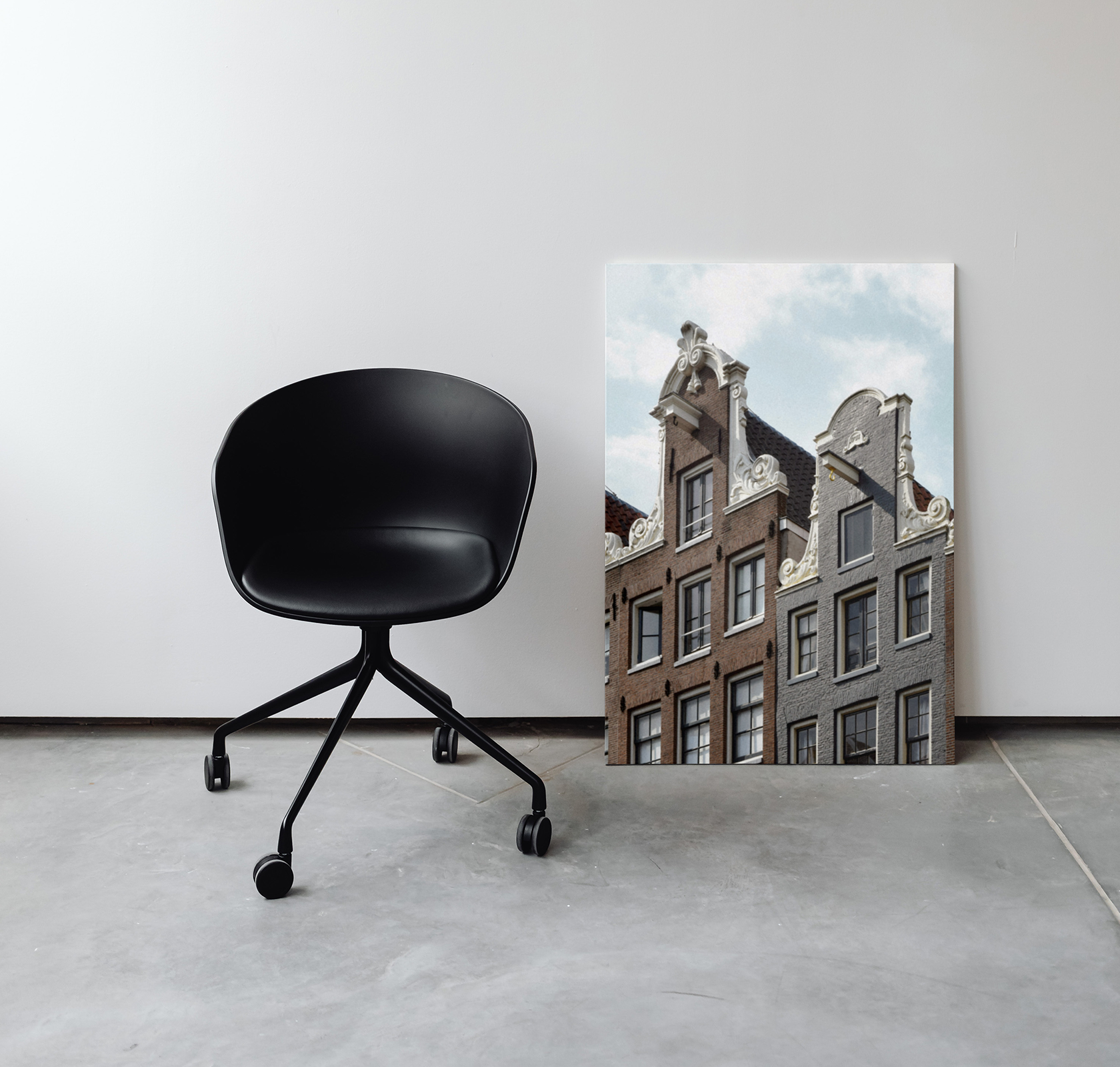 Zwarte stoel met witte muur en beton vloer met daarnaast een foto op metaal van amsterdamse panden met blauwe lucht. 