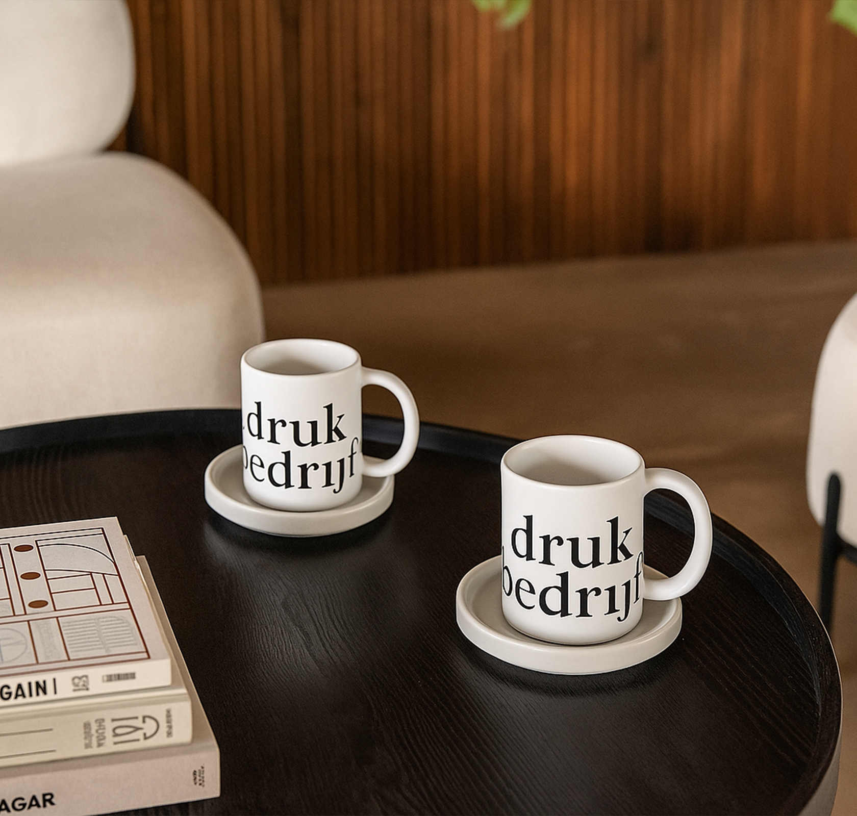 twee witte mokken bedrukt met logo op een zwart houten tafel met twee beige stoelen.
