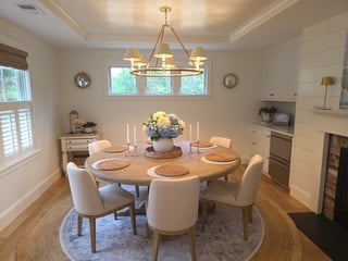 A completed dining room remodel featuring a round wood table with upholstered chairs, a brass chandelier, white walls, large windows, and built-in cabinetry.