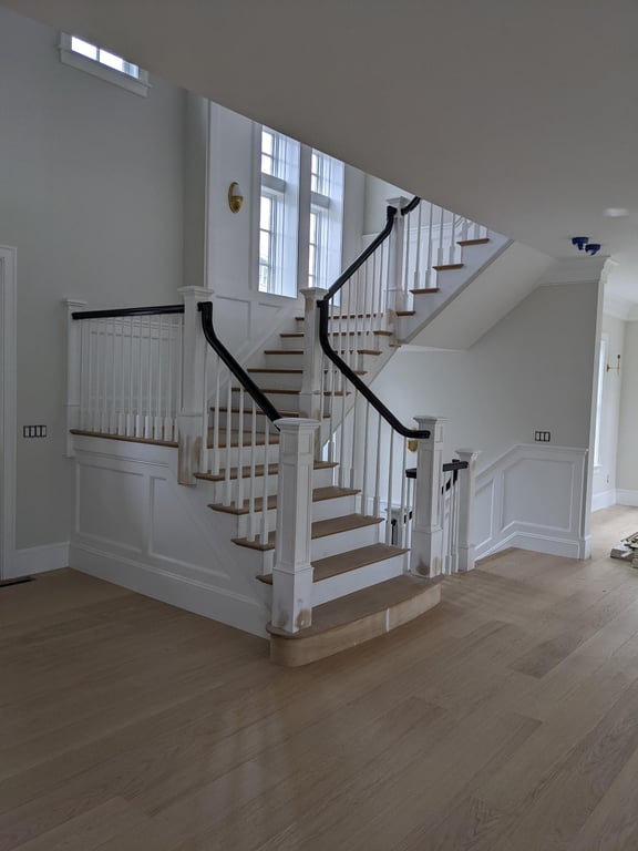 Custom staircase with white painted wooden balusters, black metal handrails, and light wood treads in a bright entry hall with large windows.