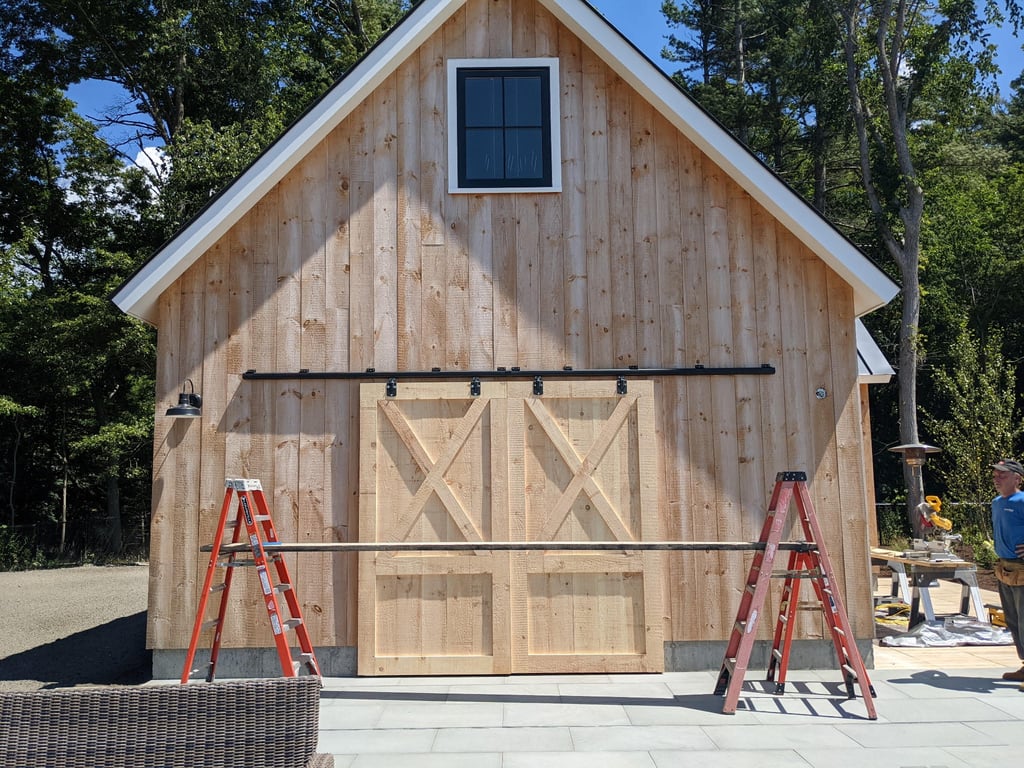 Exterior of a new pool house with natural wood siding, white trim, dark double-hung window, and large barn doors, under construction with ladders visible in front.