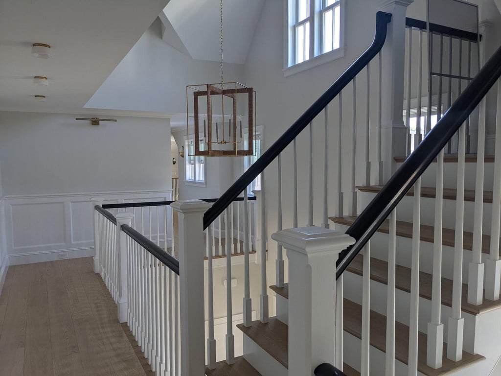 Upper hallway of a renovated home featuring a staircase with white balusters, dark wood treads, and black handrails, along with white wainscoting and recessed ceiling lights.