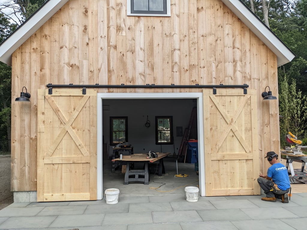 A timber-frame barn structure in Lakeville with natural wood siding, white-trimmed barn doors on sliding tracks, and a paved stone patio, shown during construction with tools and workers visible inside.