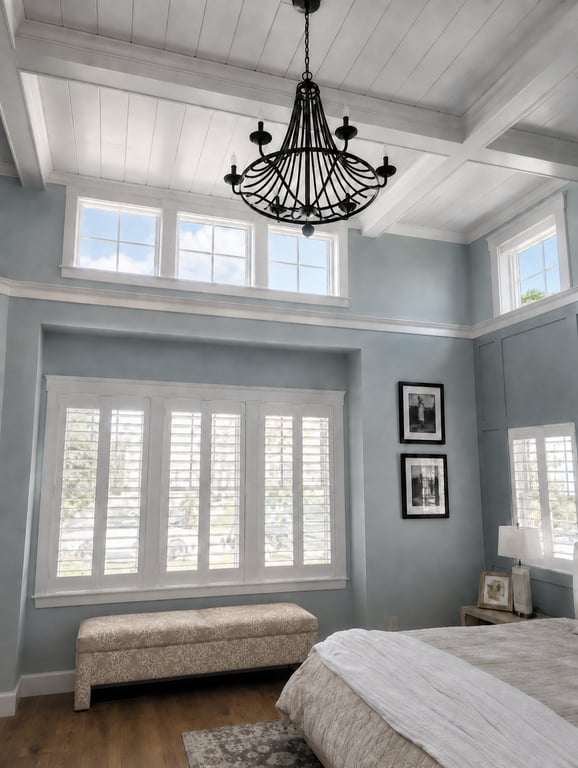A bright bedroom with blue-gray walls, white paneled ceiling with exposed beams, a black chandelier, white plantation shutters on two windows, framed artwork, an upholstered bench, and hardwood flooring.