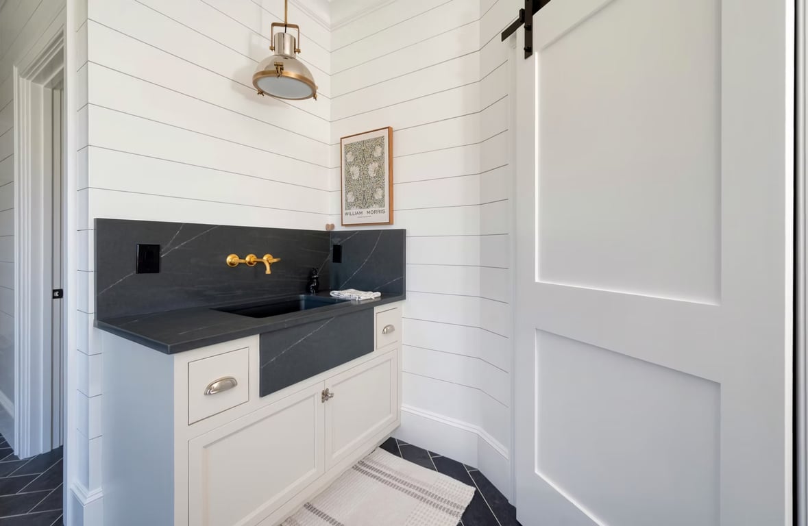 Finished bathroom with white shiplap walls, dark marble countertop and backsplash, cream cabinetry with brass hardware and faucet, black tile floor, pendant light fixture, and framed map artwork.