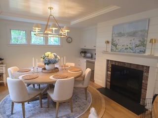 Dining room with round wood table, upholstered chairs, fireplace with white mantel, and pendant lighting fixture.