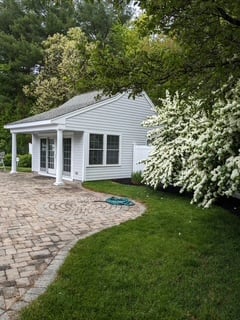 A newly constructed white cottage with covered porch and large windows, surrounded by mature landscaping and a brick paver patio.