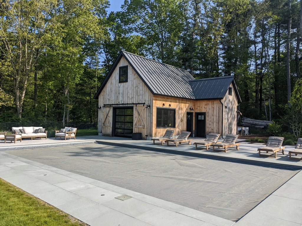 A modern pool house in Lakeville with board-and-batten siding, metal roof, and large windows, set on a concrete patio surrounded by trees.