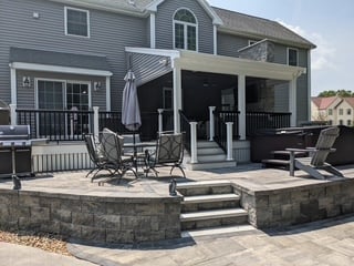 A two-story residential home with a newly constructed rear addition featuring a covered porch, deck, and stone patio with integrated seating wall.