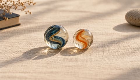 Two clear glass marbles side by side, one slightly larger, each holding a small spiral of colour, on cream linen in soft window light