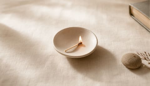 A single lit match in a small ceramic dish on cream linen — visual metaphor for hot-flash heat