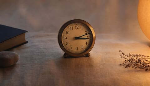 A small brass alarm clock at 3:00am beside a dimly lit lamp on cream linen
