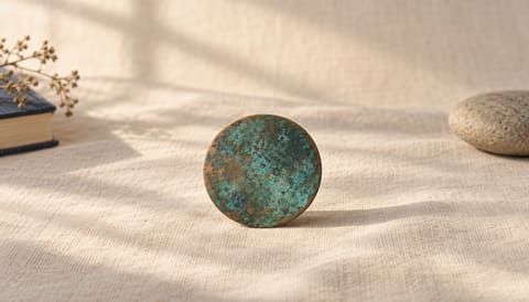 A small oxidised copper disc, green-blue patina on warm metal, on cream linen in soft window light