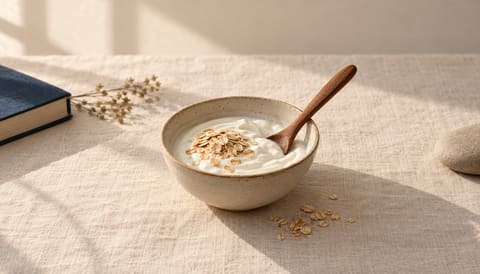 A small bowl of plain yogurt with a wooden spoon and a few oats scattered beside it, on cream linen in soft window light