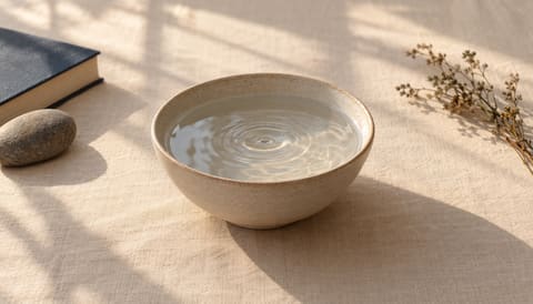 A pale ceramic bowl of still water holding a single ripple, on cream linen in soft window light