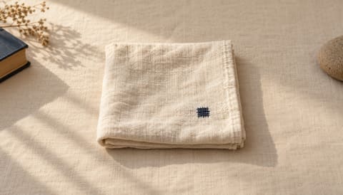 A square of folded cream linen with one darker mend stitch, on cream linen in soft window light